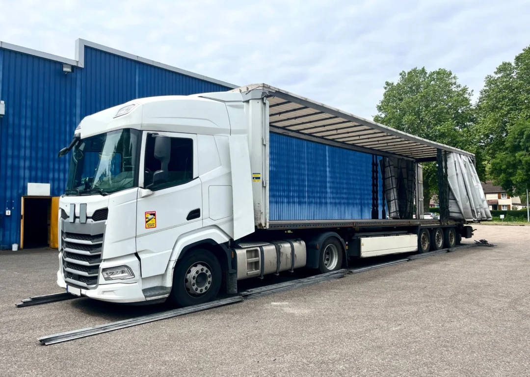 A white Scania semi-truck with a 40-foot curtain-sided trailer parked on an asphalt lot in front of a blue industrial building.