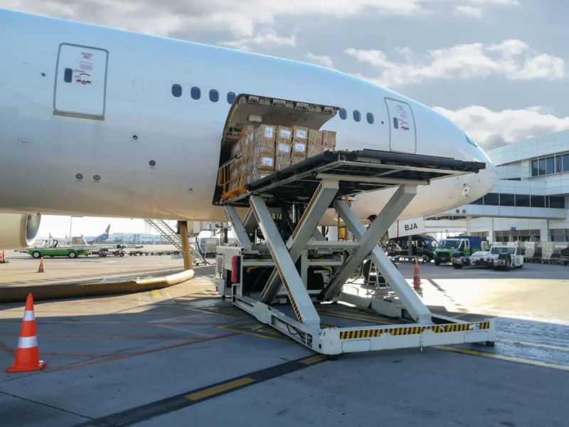 A scissor-lift cargo loader raising a large pallet of boxed air freight to the side door of a white airplane.