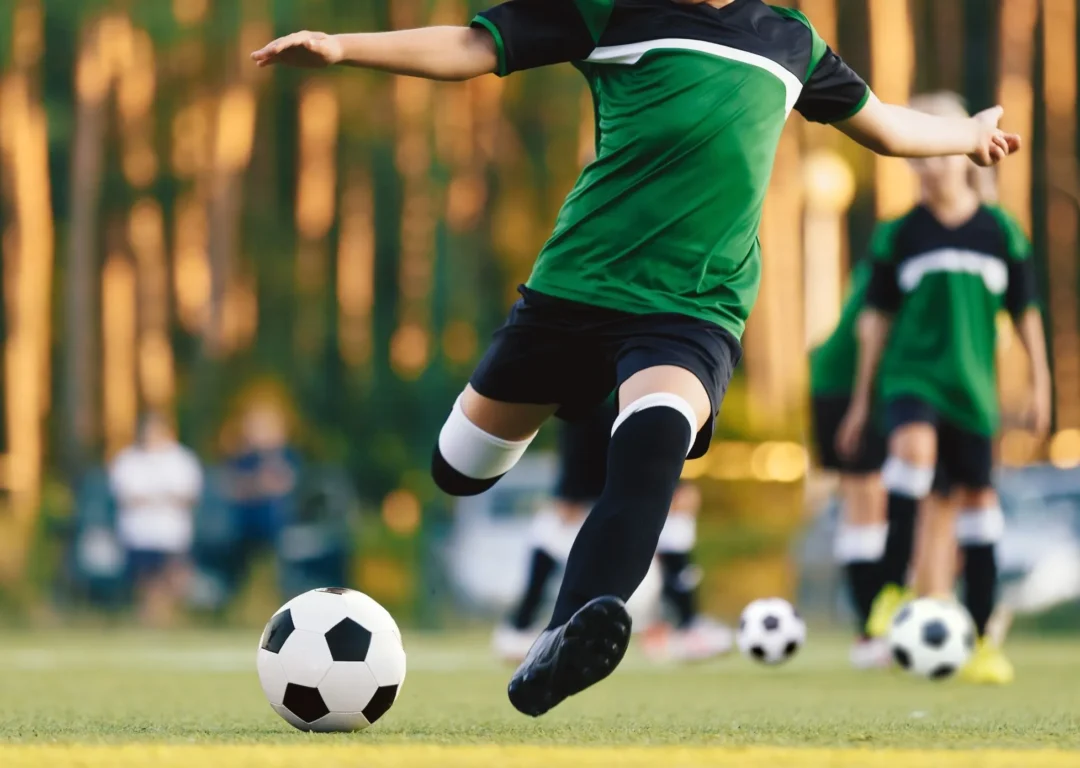 A child playing football on a grass field at sunset, wearing a green team uniform.