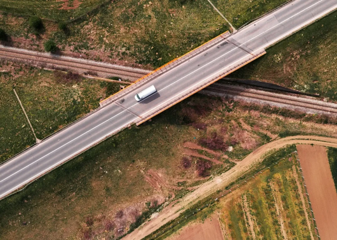 An aerial top-down view of a white delivery van driving across a bridge that spans over a railway track in a rural landscape.