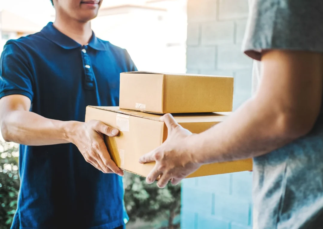 A delivery person in a blue polo shirt handing two cardboard boxes to a customer at a doorway.