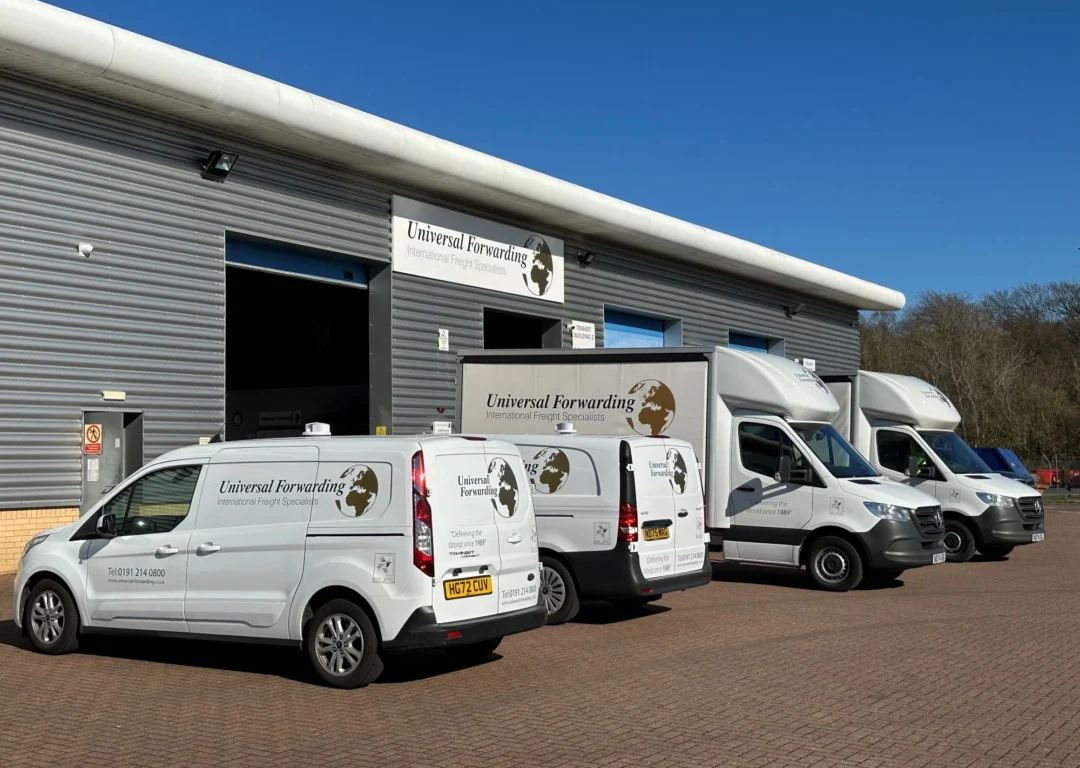 Four white Universal Forwarding delivery vans and trucks are parked in a row in front of a grey industrial warehouse under a clear blue sky.