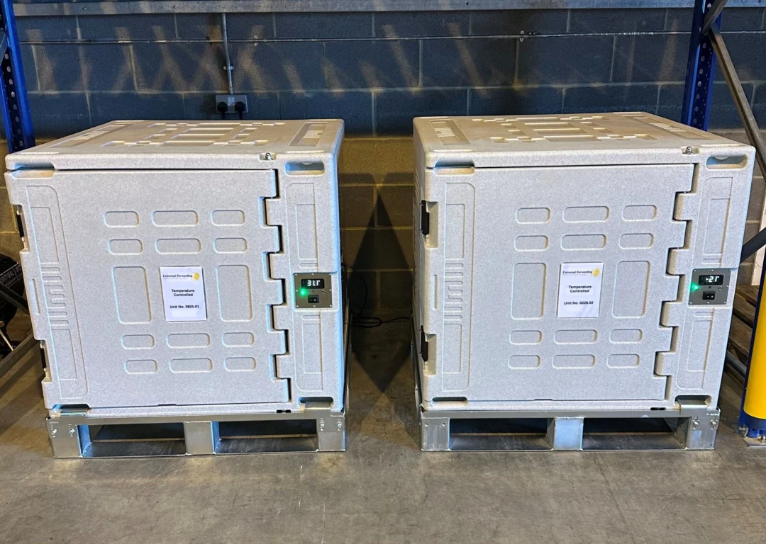 Two large, white industrial temperature-controlled shipping containers sitting side-by-side on metal pallets in a warehouse.