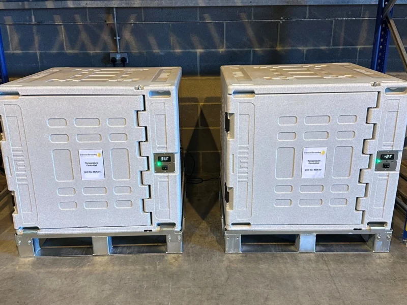 Two large, white industrial temperature-controlled shipping containers sitting side-by-side on metal pallets in a warehouse.