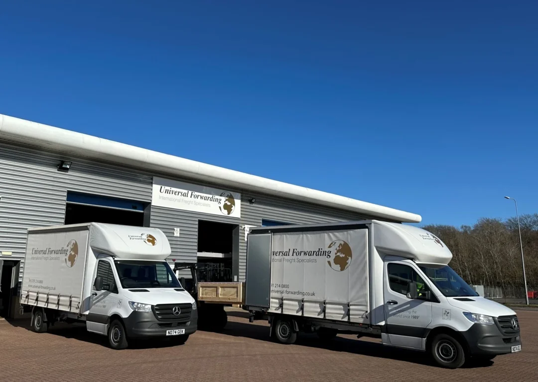 Two white Universal Forwarding delivery vans parked outside a large grey industrial warehouse under a clear blue sky.