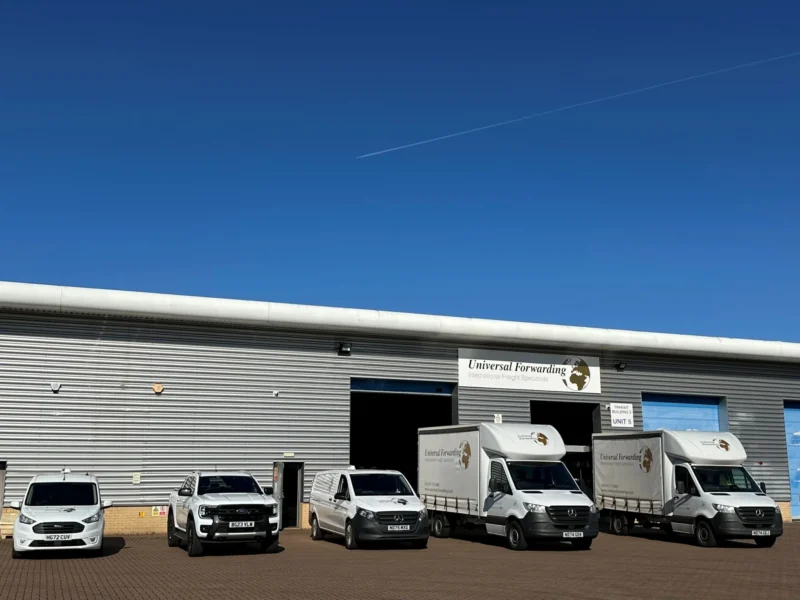 A fleet of five white Universal Forwarding delivery vehicles, ranging from vans to small trucks, parked in front of their warehouse unit under a clear blue sky.