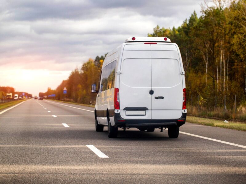 View of a white delivery van driving on a highway lined with autumn trees under a cloudy sky.