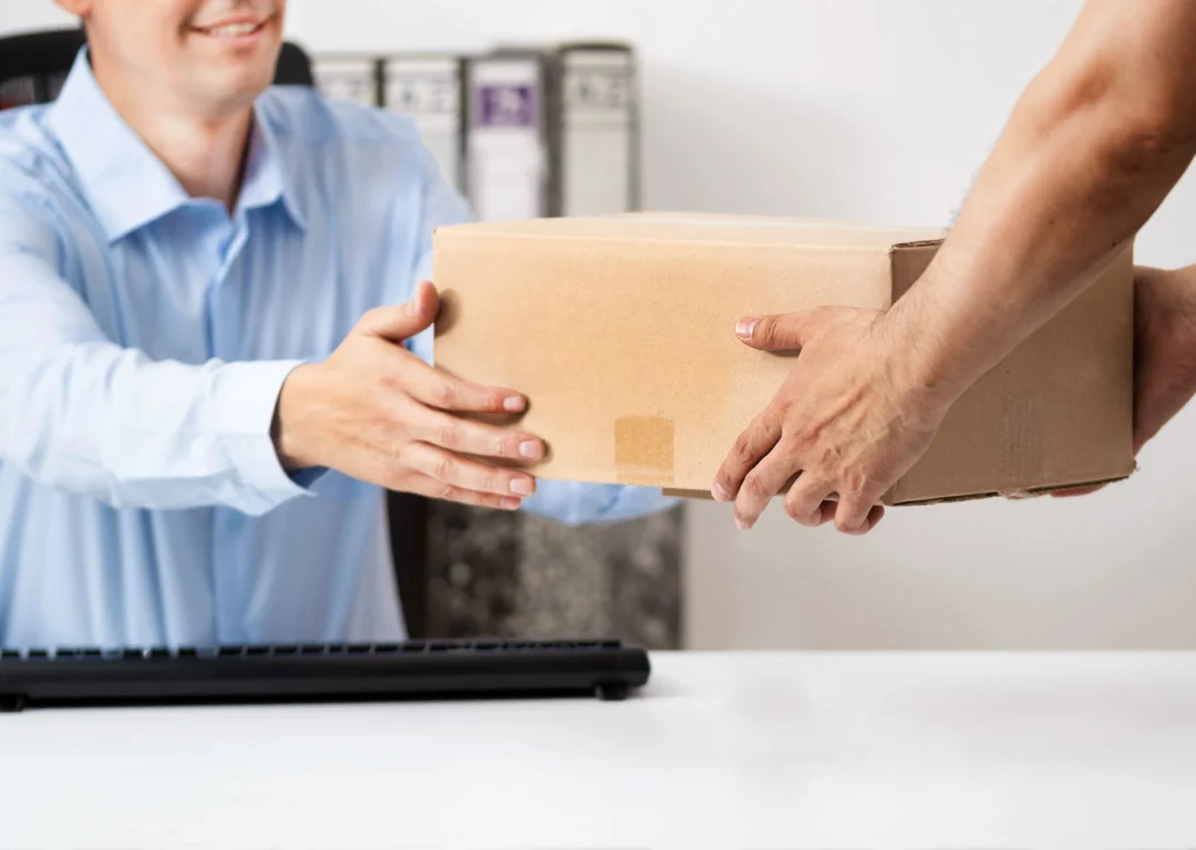 A business owner in a light blue shirt receiving a cardboard package from a delivery person.