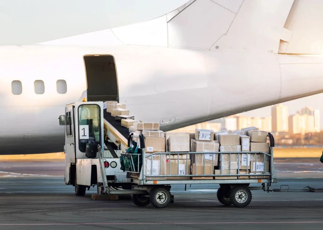 Cardboard boxes being loaded into a white cargo plane via a conveyor belt loader on an airport tarmac.