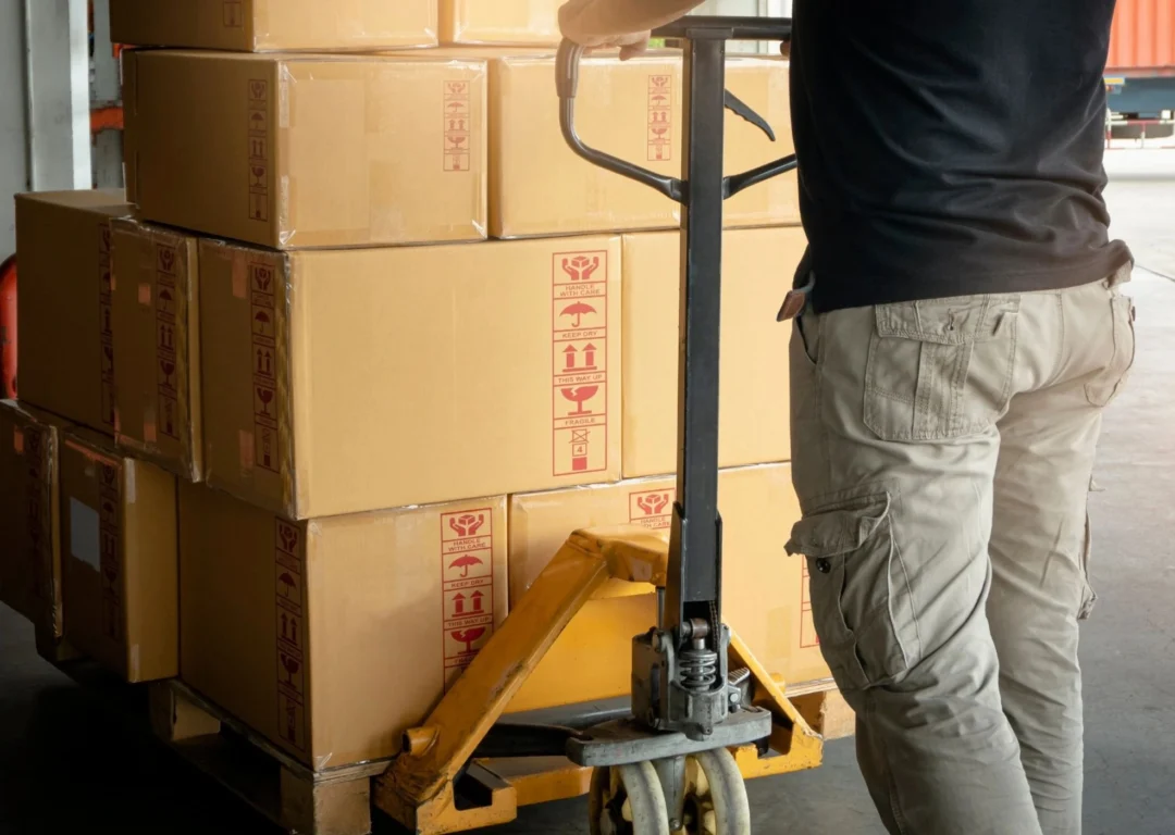 A warehouse worker uses a yellow pallet jack to move a stack of brown cardboard boxes marked with 'handle with care' symbols.