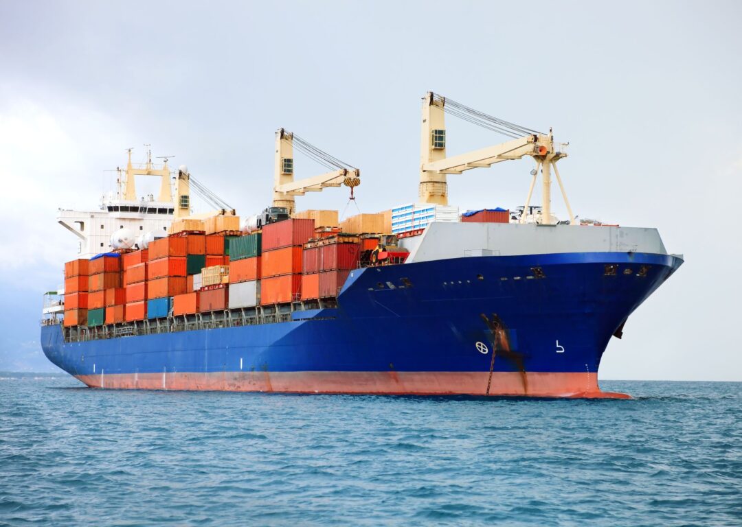 A large blue container ship with onboard cranes, heavily loaded with orange and red shipping containers, sailing on the ocean.