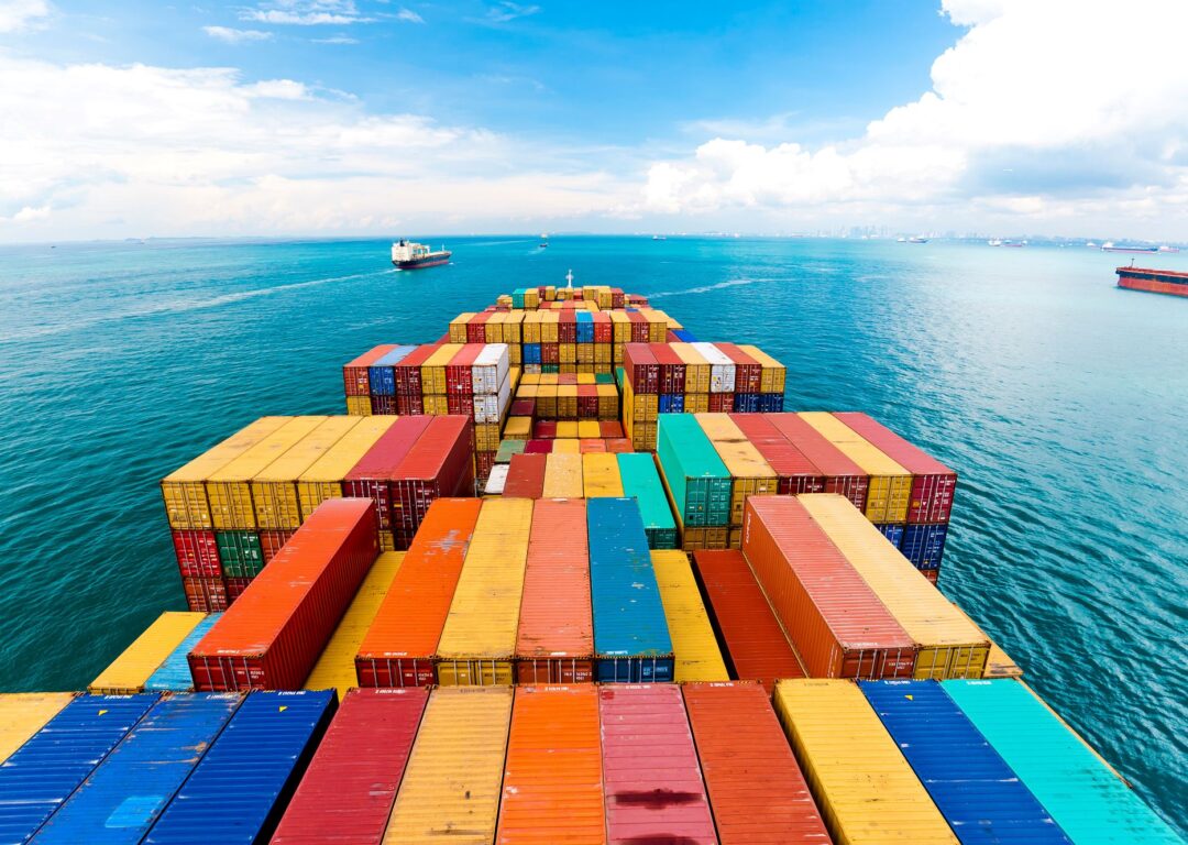 The deck of a container ship, looking out over rows of colourful shipping containers toward a sunny blue ocean horizon with other vessels in the distance.