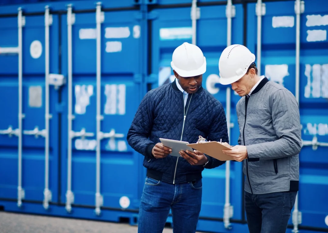 Two logistics workers in white hard hats looking at a tablet and clipboard in front of blue shipping containers.
