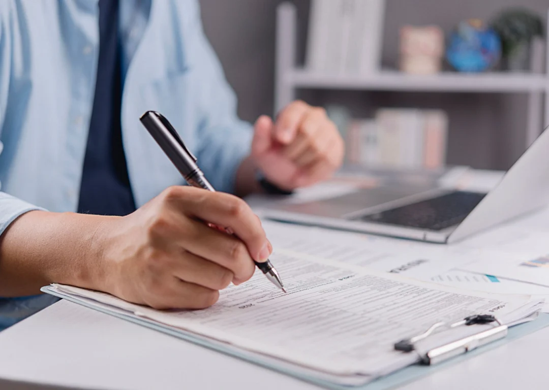 A person's hand holding a pen, filling out a shipment insurance form on a clipboard next to a laptop.