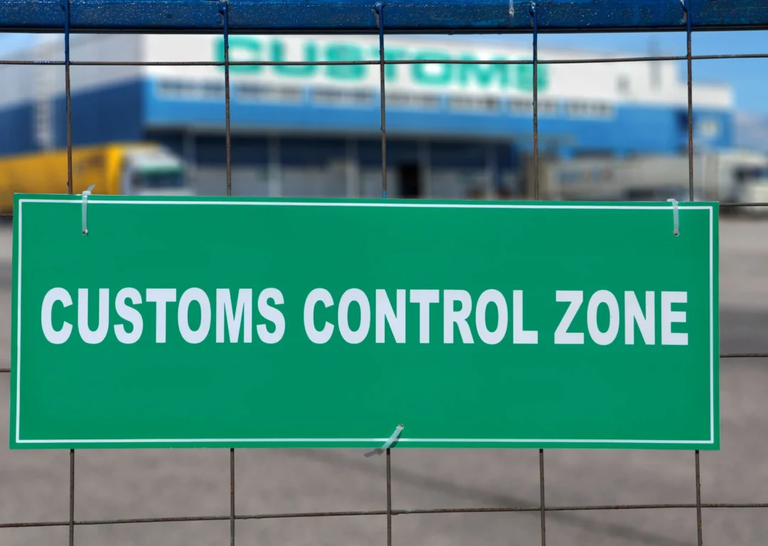 A green 'customs control zone' sign attached to a wire fence, with a blurred logistics building and cargo trucks in the background.