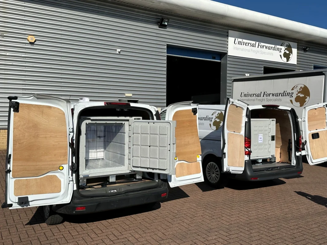 White Universal Forwarding vans with open rear doors, showing temperature-controlled storage units loaded inside, parked in front of a warehouse.