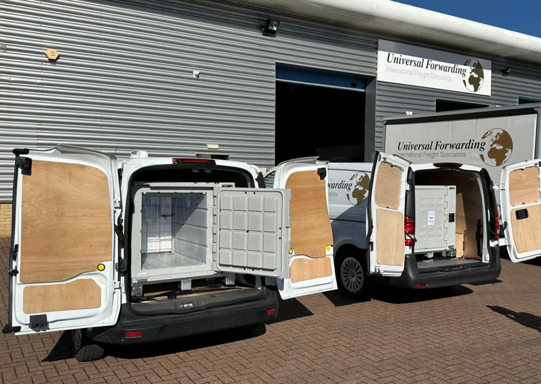White Universal Forwarding vans with open rear doors, showing temperature-controlled storage units loaded inside, parked in front of a warehouse.