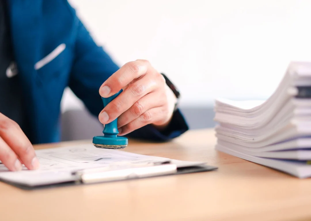 A person in a blue blazer using a blue rubber stamp on a document resting on a clipboard, with a large stack of paperwork nearby.