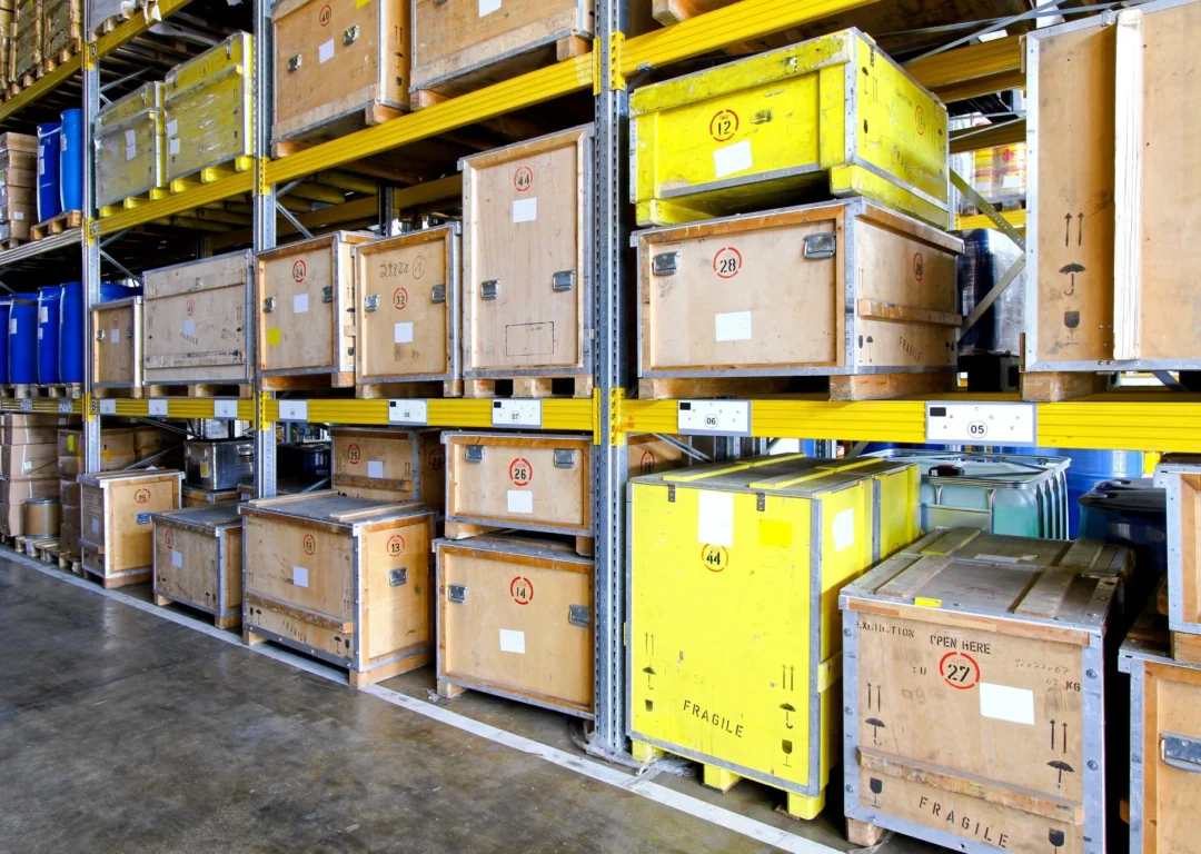 Wooden crates and blue barrels stored on industrial yellow metal shelving in a warehouse.