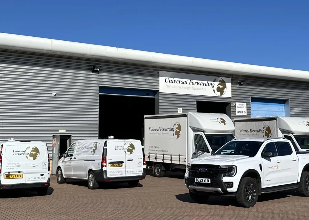 A fleet of five white Universal Forwarding vehicles, including vans, a pickup truck, and a small curtain-side truck, parked outside a grey industrial warehouse.